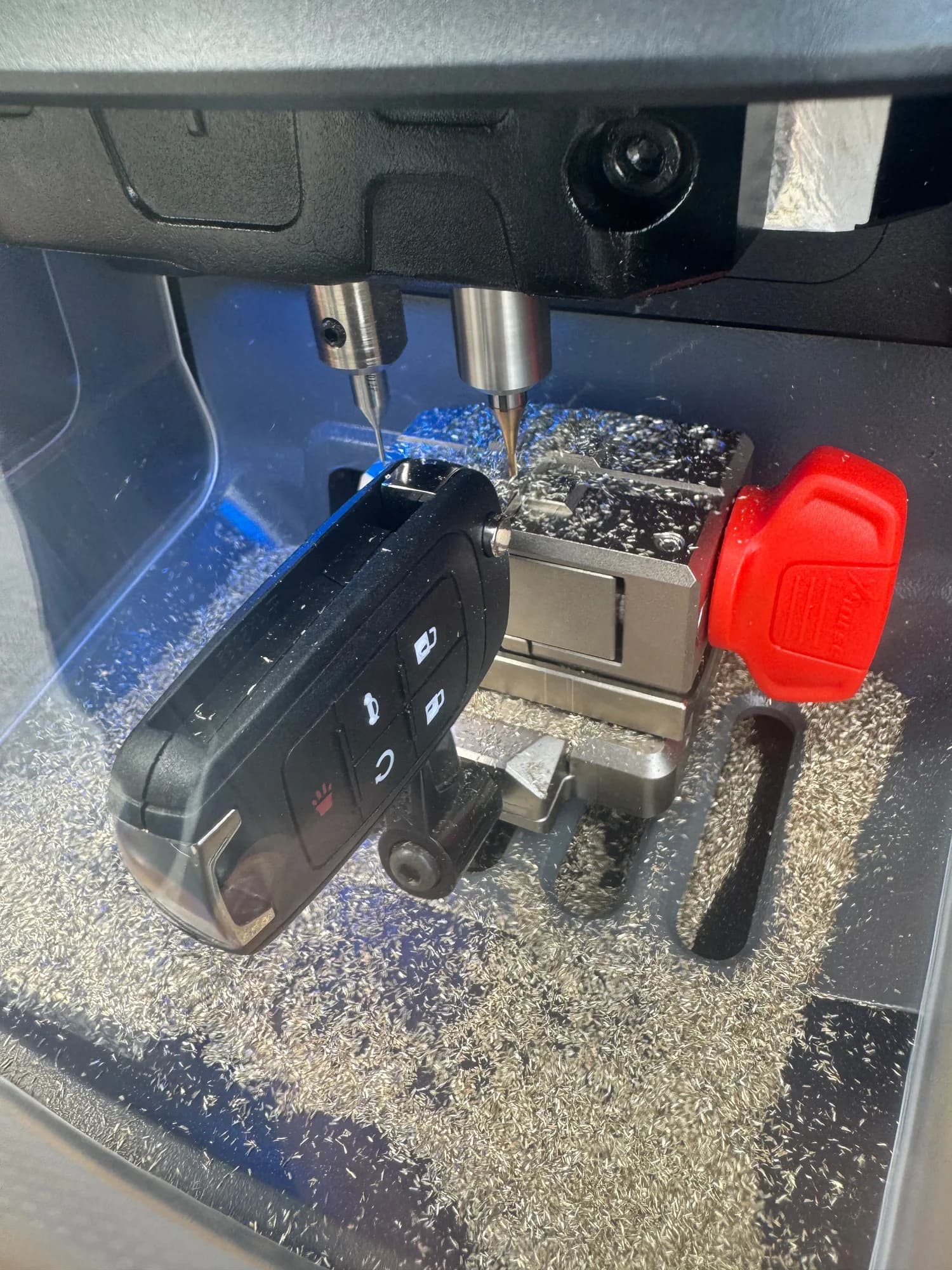 A car key blank clamped in the jaw of a key cutting machine with the cutter above it and metal shavings across the bed, the finished flip key resting alongside